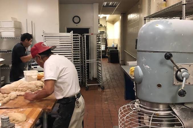 Two bakers work dough beside racks of trays and a large mixer.