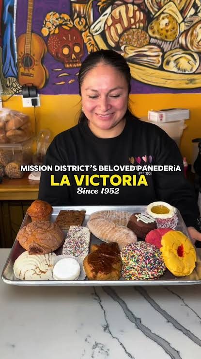 A team member holds a tray of pan dulce in front of a colorful mural.