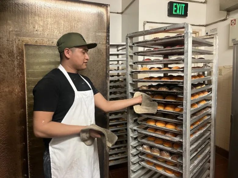 A baker handles a tray on a rack of freshly baked breads near an oven.