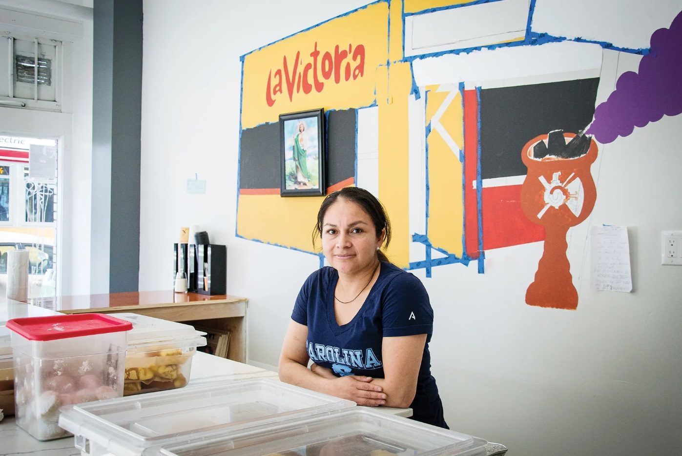 A bakery staff member sits at the counter in front of a painted La Victoria mural.